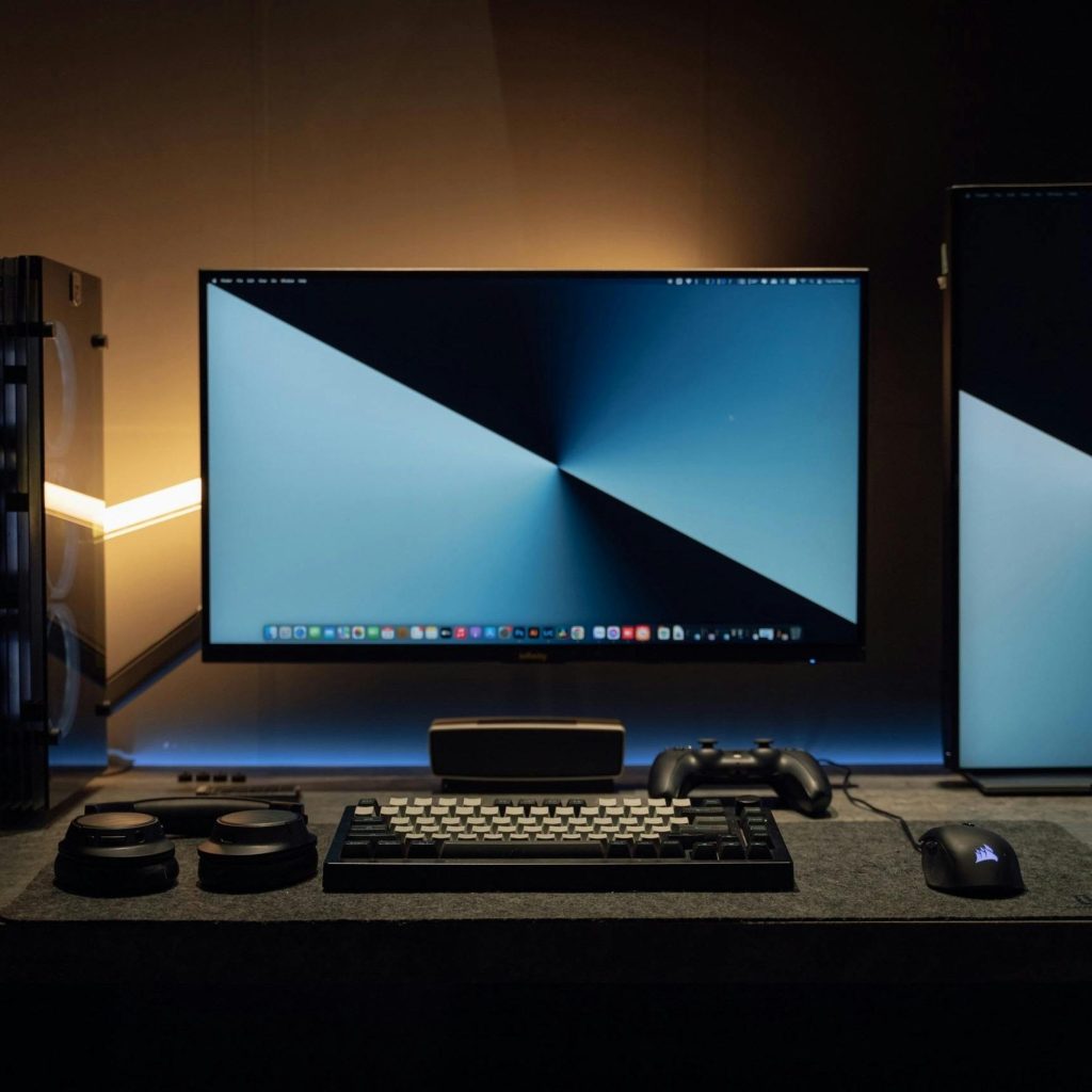 A dimly lit modern computer desk setup featuring dual monitors, a keyboard, and gaming accessories.